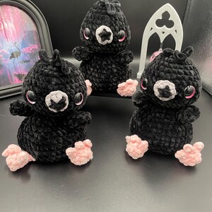Peut inclure: Trois peluches d'oiseaux noirs au crochet, faites à la main, avec des pieds et des accents roses. Chaque oiseau a de grands yeux ronds avec des reflets roses et un visage blanc et gris. Les peluches sont disposées sur une surface sombre.