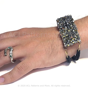 Puede incluir: Una pulsera elástica negra con un dije rectangular de cuentas plateadas y doradas. La pulsera está en la muñeca de una persona. La persona también lleva un anillo de plata con piedras verdes.