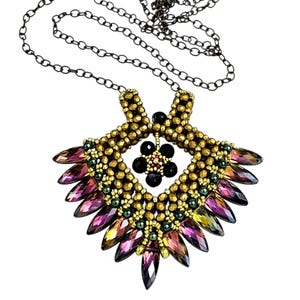 May include: A handmade necklace with a geometric pendant. The pendant features gold-coloured beads, iridescent purple and green crystals, and black accents. The necklace has a dark chain.