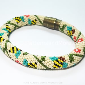 May include: Cream-coloured beaded bracelet with a floral and geometric pattern. The bracelet features a repeating design of green vines, red, yellow, and turquoise flowers, and black and yellow bee-like shapes. The clasp is a bronze-coloured magnetic closure.