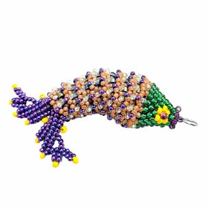 Peut inclure: Un charme de poisson en perles avec une queue violette, un corps brun et blanc et une tête verte avec une fleur jaune.