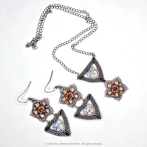 Puede incluir: Un collar de cadena plateada con un colgante triangular que presenta una cuenta iridiscente rosa y morada. El colgante está rodeado de cuentas plateadas y tiene dos grupos de cuentas más pequeños en forma de estrella que cuelgan de él. También se muestran pendientes a juego con el mismo diseño.