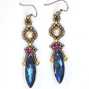 May include: A pair of handcrafted earrings with a dark gunmetal finish. Each earring features a faceted, deep blue teardrop-shaped crystal, accented with pink and iridescent beads. The earrings are suspended from a silver-tone fish hook. © 2021 ACL Patterns and More. All rights reserved.