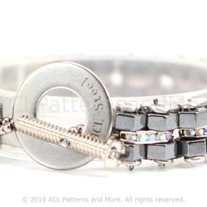 May include: A silver bracelet with a black and silver chain and clear crystals. The bracelet has a silver clasp with the word "Steel" engraved on it.