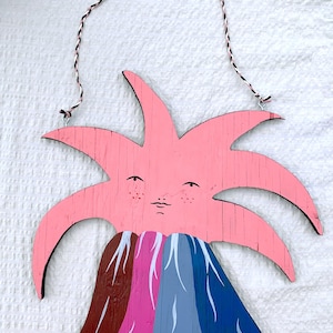 May include: A whimsical wooden wall hanging featuring a pink sun with a smiling face and a colorful volcano. The volcano has brown, pink, gray, and blue layers with white accents. A black and white string is attached for hanging.