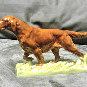 May include: A ceramic figurine of a reddish-brown dog, possibly an Irish Setter, in a walking pose. The dog is on a light green base with a textured design. The figurine has detailed features and a glossy finish, showcasing a classic decorative item.