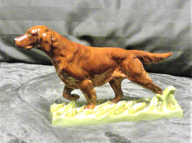May include: A ceramic figurine of a reddish-brown dog, possibly an Irish Setter, in a walking pose. The dog is on a light green base with a textured design. The figurine has detailed features and a glossy finish, showcasing a classic decorative item.