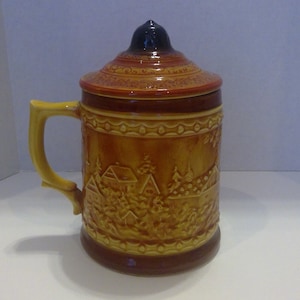 May include: A ceramic beer stein with a lid, featuring a brown and yellow glaze with a scene of a village. The stein has a handle and a decorative design around the top.