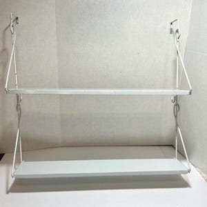 May include: A white, two-tiered wall-mounted shelf. The shelves are rectangular and supported by white metal brackets that attach to the wall. The shelf is designed for displaying items or storage.