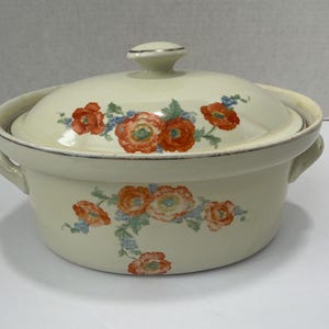 May include: Cream-colored ceramic casserole dish with lid. The dish features a floral design with orange and red flowers, green leaves, and blue accents. It has two handles and a silver rim.