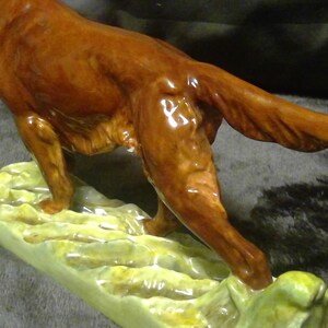May include: A glossy brown ceramic dog figurine, depicted mid-stride with its tail extended. The dog stands on a light green base with a textured, organic design. This decorative figurine is a detailed representation of a dog, suitable for display.