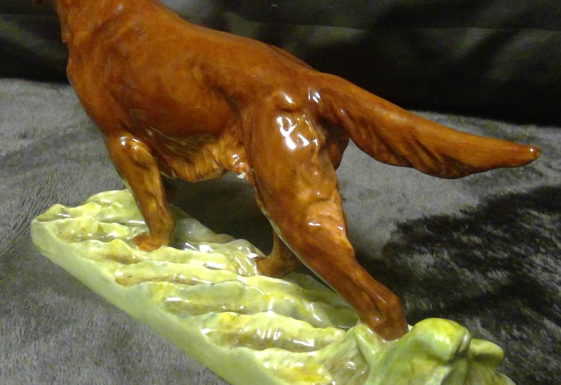 May include: A glossy brown ceramic dog figurine, depicted mid-stride with its tail extended. The dog stands on a light green base with a textured, organic design. This decorative figurine is a detailed representation of a dog, suitable for display.