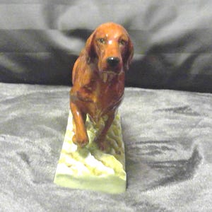 May include: A ceramic figurine of a reddish-brown dog, possibly a setter, walking forward. The dog is on a rectangular base with a textured, off-white surface. The figurine has a glossy finish.