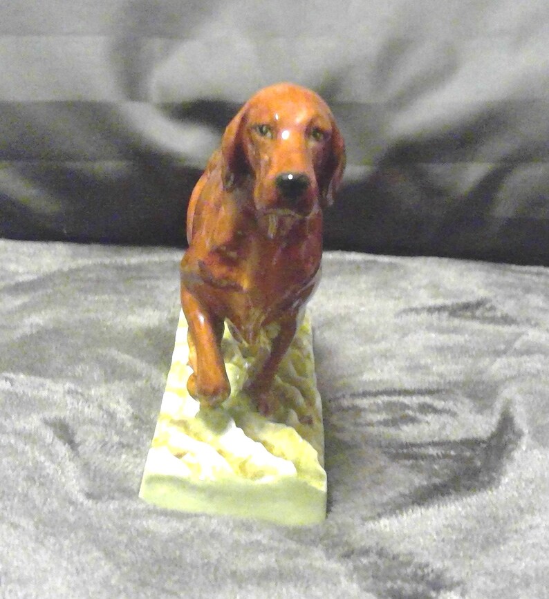 May include: A ceramic figurine of a reddish-brown dog, possibly a setter, walking forward. The dog is on a rectangular base with a textured, off-white surface. The figurine has a glossy finish.