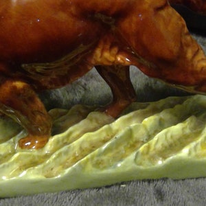 May include: A ceramic dog figurine, possibly a hunting breed, in a running stance. The dog is a glossy, rich brown with detailed legs and paws. It stands on a base of stylized, textured, light green and yellow grass.