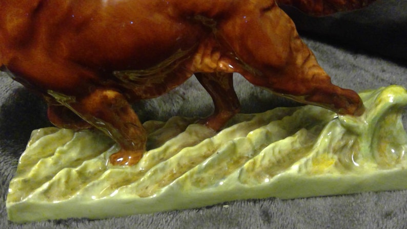 May include: A ceramic dog figurine, possibly a hunting breed, in a running stance. The dog is a glossy, rich brown with detailed legs and paws. It stands on a base of stylized, textured, light green and yellow grass.