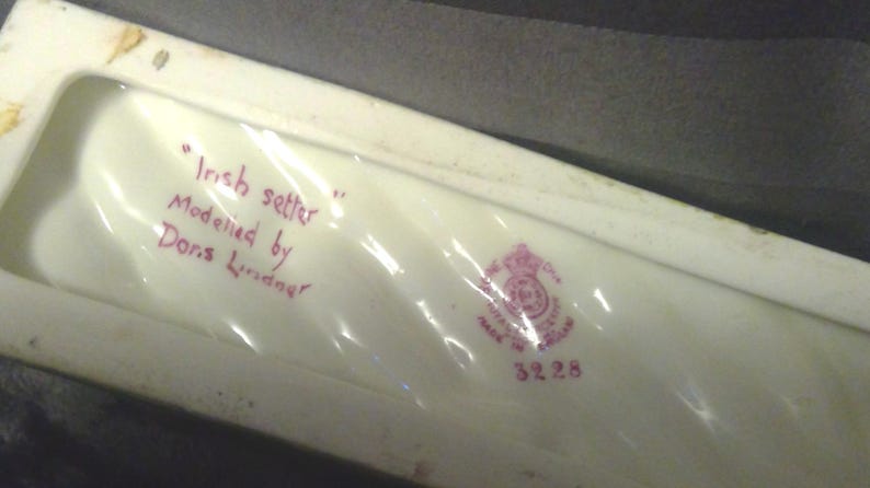 May include: An off-white ceramic piece with the text "Irish setter" and "Modelled by Doris Lindner" in red ink. A pink crest and the number 3228 are also visible. The piece is rectangular.