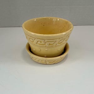 May include: A light yellow ceramic planter with a matching saucer. The planter has a rounded shape with a decorative geometric pattern around the rim and a series of small holes. The saucer has a similar design.