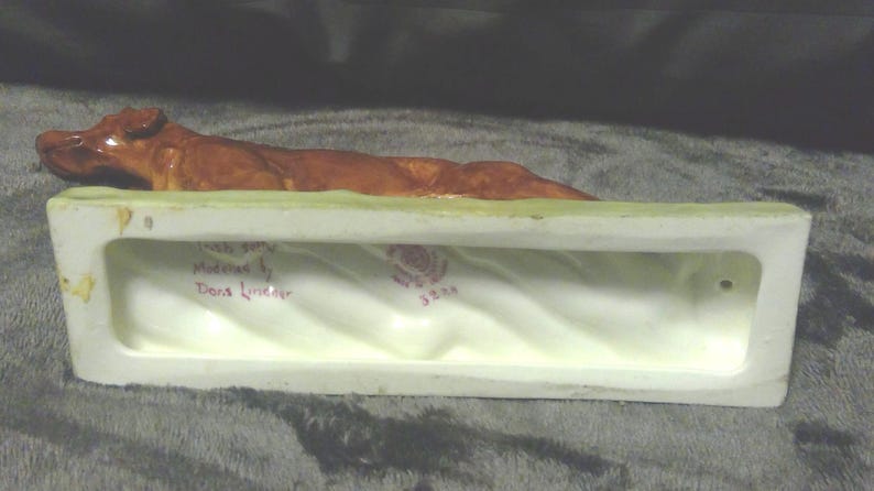 May include: A ceramic ornament featuring a brown dog resting on a rectangular, white base with a green top. The base has a wavy interior and text that reads "Irish Setter" and "modelled by Dons Linley".