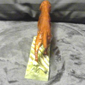 May include: A carved wooden sculpture of a horse, standing upright on a rectangular base. The horse is a reddish-brown colour, with detailed carving. The base is light green with yellow and white stripes.
