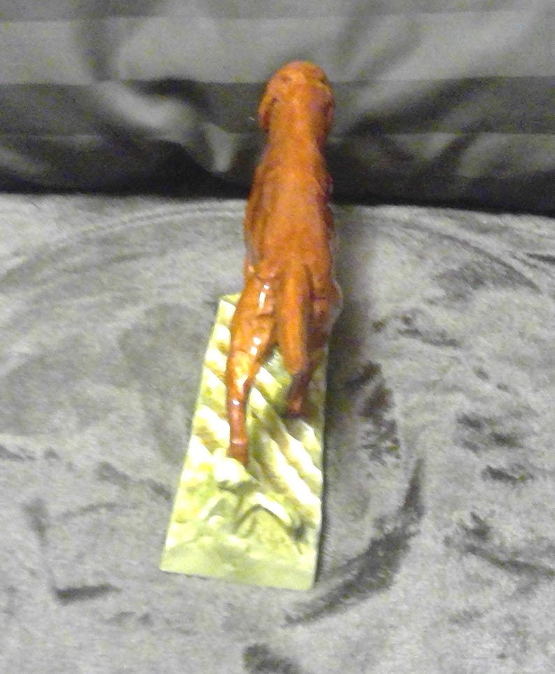 May include: A carved wooden sculpture of a horse, standing upright on a rectangular base. The horse is a reddish-brown colour, with detailed carving. The base is light green with yellow and white stripes.