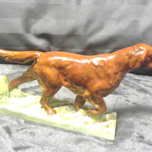 May include: A ceramic dog figurine in a walking pose. The dog is a reddish-brown colour with a glossy finish. It is mounted on a rectangular base with a light green and yellow colour.