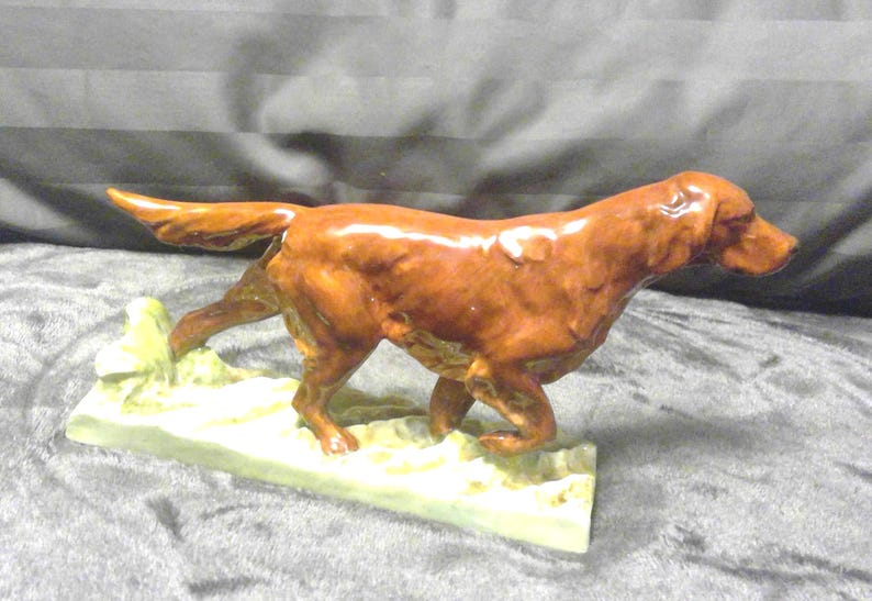 May include: A ceramic dog figurine in a walking pose. The dog is a reddish-brown colour with a glossy finish. It is mounted on a rectangular base with a light green and yellow colour.