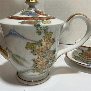 May include: White porcelain Japanese tea set with gold accents. The teapot features a lid with a gold finial and is decorated with floral and mountain designs. A teacup and saucer are also visible.