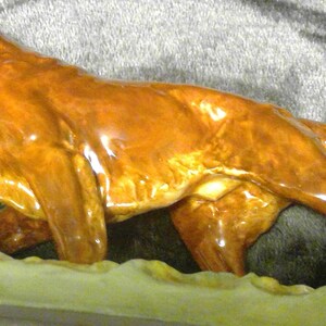 May include: A ceramic dog figurine in a running pose, with a glossy, reddish-brown finish. The dog is depicted with detailed features and is set against a neutral background. The figurine is likely a decorative item.