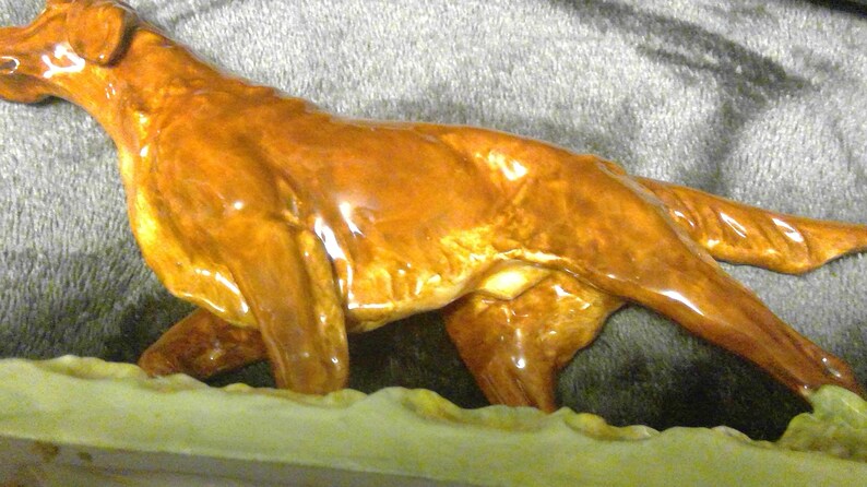 May include: A ceramic dog figurine in a running pose, with a glossy, reddish-brown finish. The dog is depicted with detailed features and is set against a neutral background. The figurine is likely a decorative item.