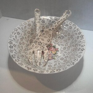 May include: A white ceramic salad bowl with a floral pattern in silver and gold. The bowl is paired with two matching serving forks.