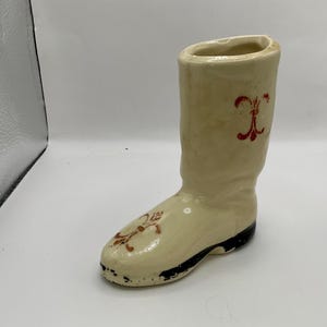 May include: A cream-colored ceramic boot-shaped vase with a black base and red floral accents. The vase has a tall, cylindrical shaft and a detailed design on the front. The boot is approximately 7 inches tall.
