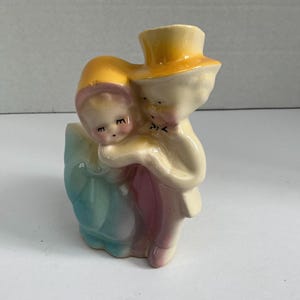 May include: A vintage ceramic figurine depicting a couple embracing. The man wears a yellow top hat and coat, while the woman is in a blue and pink dress. The figurine is painted in pastel colors and has a glossy finish.