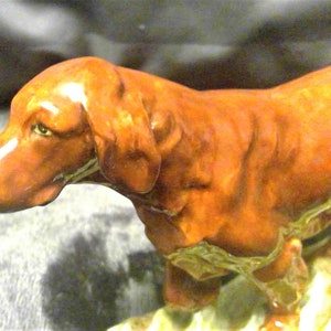May include: A ceramic figurine of a brown dog with a long snout and floppy ears. The dog is standing on a patch of green and brown foliage. The figurine has a glossy finish and is likely a decorative item.