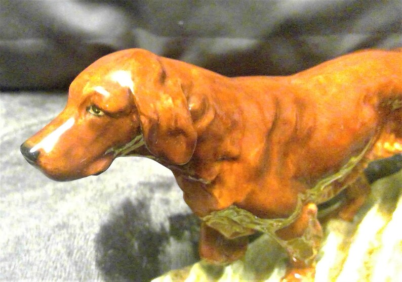 May include: A ceramic figurine of a brown dog with a long snout and floppy ears. The dog is standing on a patch of green and brown foliage. The figurine has a glossy finish and is likely a decorative item.