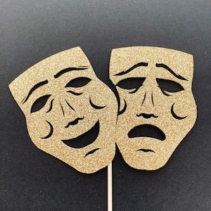 May include: Two gold glitter paper masks, one with a smiling face and one with a frowning face, attached to a wooden stick. The masks are a traditional symbol of comedy and tragedy.