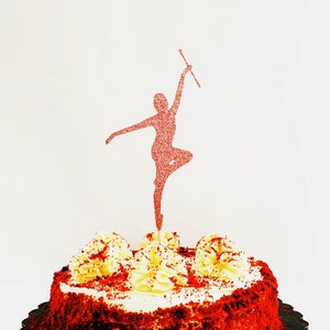 May include: Red velvet cake with white frosting and a red glitter silhouette of a figure holding a baton on top. The cake is on a black scalloped cake board. The text "Patty Antle's Prettys" is on the bottom of the image.