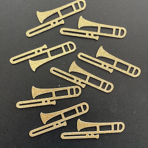 May include: A collection of ten gold glitter paper cutouts of trombones. The trombones are arranged in a random pattern on a black background. The text "Patty Antle's Prettys" is at the bottom of the image.
