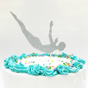 May include: A silver glitter cake topper shaped like a person diving, positioned on a white frosted cake with turquoise blue piped frosting borders and colorful sprinkles. The text Patty Antle's Prettys pattyantle.com is visible in the background.