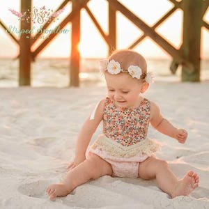 May include: A baby girl wearing a floral print romper with a white fringe trim and a flower crown. The baby is sitting on a sandy beach.