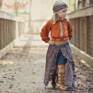 May include: A young girl wearing a rust-colored peasant top with long sleeves, a floral print skirt, and gladiator sandals. The skirt has a high-low hemline. A matching floral headband completes the outfit. The image is set outdoors on a wooden bridge.