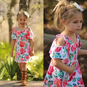 May include: A young girl wearing a white dress with a floral print and cold shoulder design. The dress has pink, red, and green flowers. She is wearing brown sandals and has a white flower in her hair.