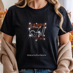 May include: A black short-sleeved t-shirt featuring the text "HAPPY FALL Y'ALL" in orange and white rhinestones with a starburst effect. Below the main graphic, the text "time • truth • hearts" is printed in small white sans-serif letters.