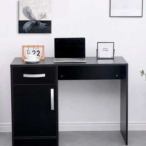 May include: A black desk with a drawer and a cabinet door. The desk has a black top and a white wall behind it. There is a laptop computer, a calendar, and a picture frame on the desk.
