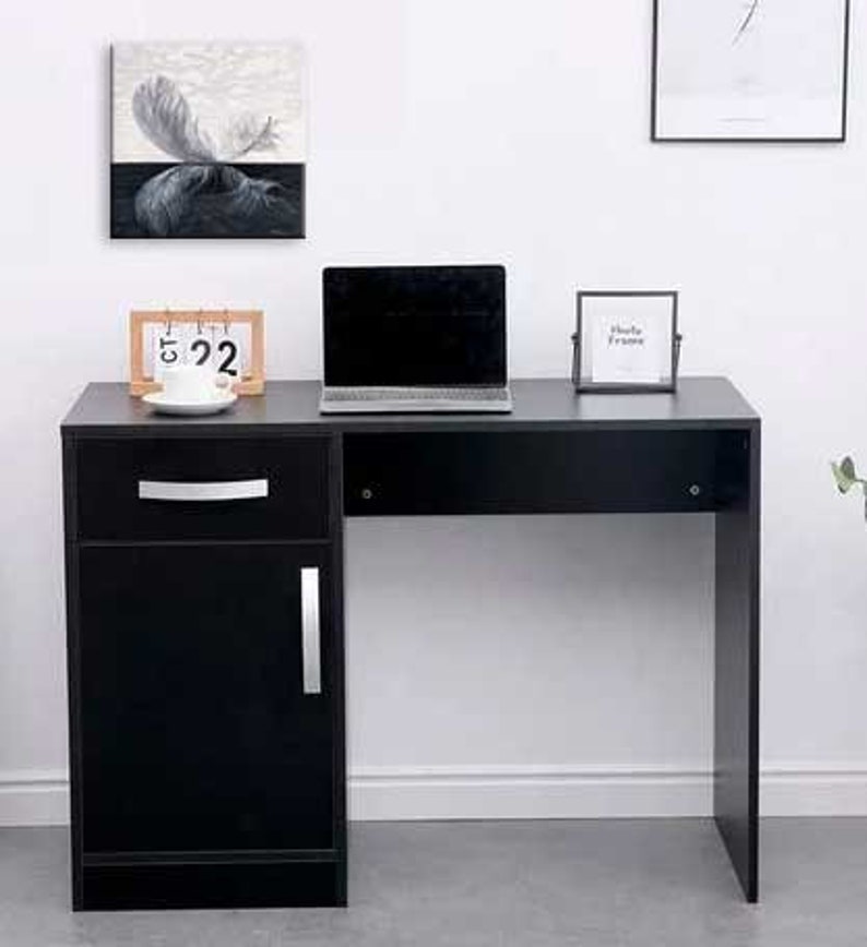 May include: A black desk with a drawer and a cabinet door. The desk has a black top and a white wall behind it. There is a laptop computer, a calendar, and a picture frame on the desk.