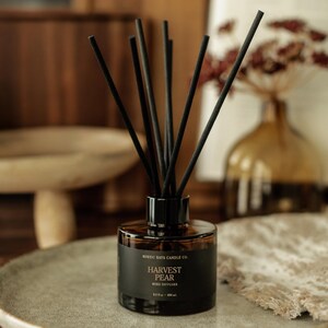 May include: A brown glass reed diffuser with black reeds. The label reads "Harvest Pear Reed Diffuser" from Rustic Rays Candle Co. The diffuser sits on a gray plate. The bottle contains 3.5 fl oz (104 ml).