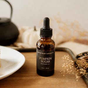 May include: A brown glass dropper bottle with a black cap and a black label. The label features the text Rustic Rays Candle Co. Pumpkin Sugar Diffuser Oil 1 fl oz 30 mL. The bottle contains a liquid and is topped with a black rubber dropper bulb.