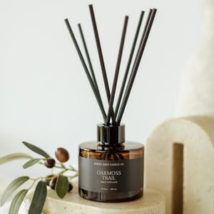 May include: A brown glass reed diffuser with black reeds. The label reads "OAKMOSS TRAIL REED DIFFUSER" and "6.8 fl oz - 104 ml". The diffuser is on a stone surface with a sprig of olives.