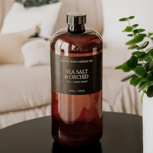 May include: A brown glass bottle of air and linen spray with a black cap. The label reads "Sea Salt & Orchid" and "Rustic Rays Candle Co." The bottle contains 16 fl oz (473 mL) of product. The bottle is on a black surface.