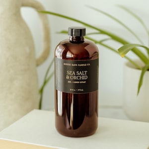 May include: A brown plastic bottle with a black cap and a black label featuring the text Rustic Rays Candle Co. Sea Salt & Orchid Air + Linen Spray 16 fl oz 473 mL. The bottle has a cylindrical shape and a smooth, reflective surface finish.
